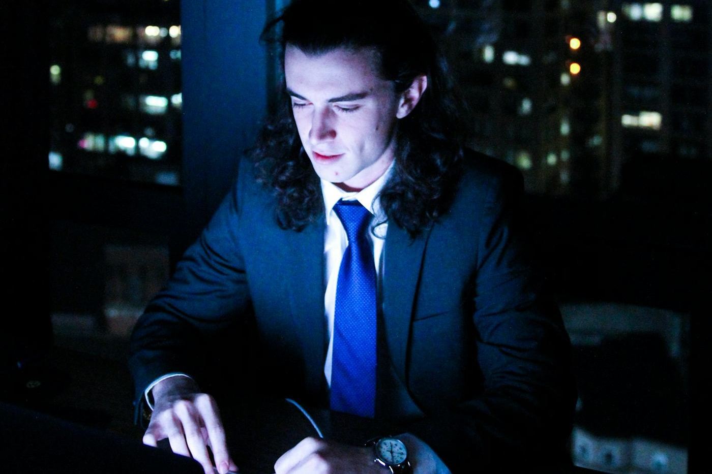 Executivo de terno trabalhando de madrugada, iluminado pela luz azul do notebook, com a cidade ao fundo